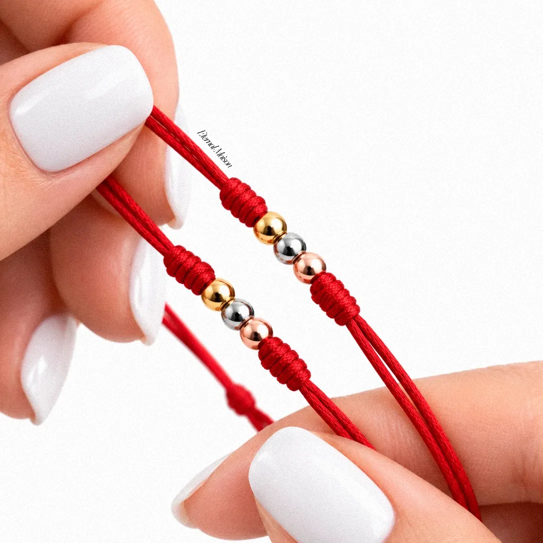 Manos sosteniendo dos pulseras de cordón rojo con balines en oro amarillo, blanco y rosado.